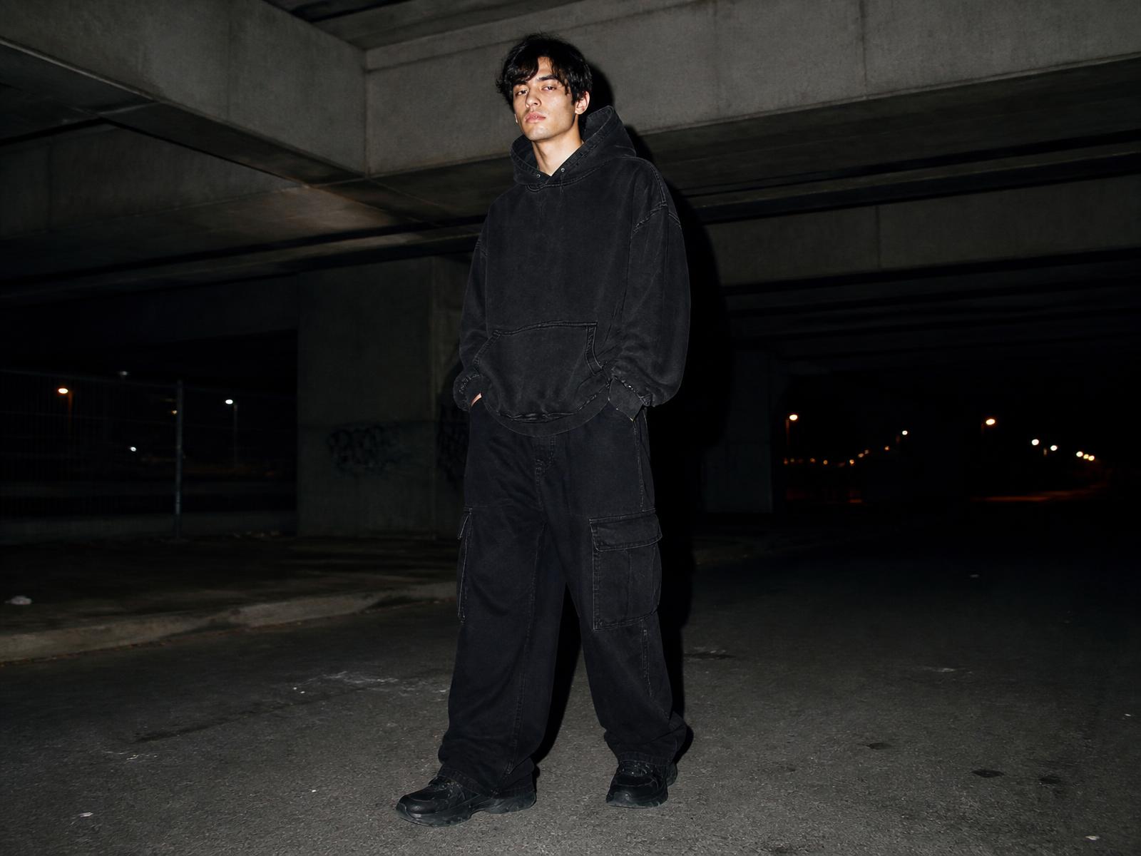 Model in a washed black hoodie and wide cargos under a concrete underpass at dusk